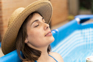 Young woman wearing sun hat while relaxing in inflatable swimming pool at yard - AFVF06823