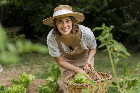 Lächelnde junge Frau mit Hut sammelt Gemüse in einem Weidenkorb im Garten, lizenzfreies Stockfoto