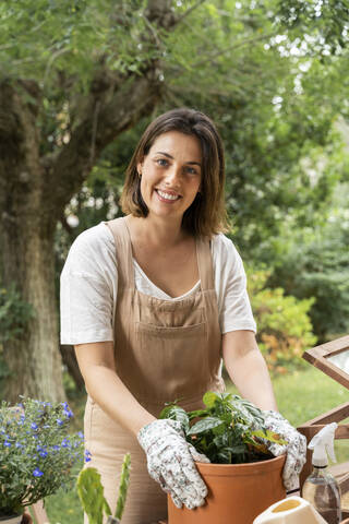 Smiling young woman holding potted plant while standing against tree in yard during curfew stock photo