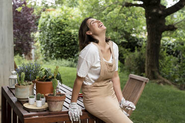 Cheerful young woman laughing while sitting on table in yard during curfew - AFVF06840