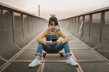 Stylischer Teenager in Jeans-Overall, der auf einem Skateboard sitzt und ein Mobiltelefon auf einem Fußweg in der Stadt benutzt - ADSF04864