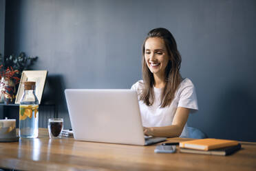 Smiling young woman using laptop on desk against wall in home office - BSZF01572