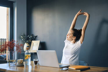 Tired young woman stretching arms while sitting at desk in home office - BSZF01575