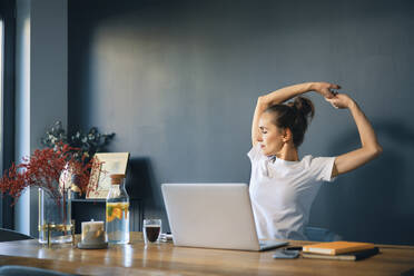 Tired businesswoman stretching arms while sitting at desk in home office - BSZF01576