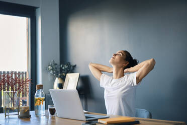 Tired businesswoman with hands behind head sitting at desk in home office - BSZF01577