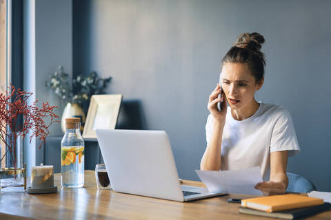 Young woman holding document talking over smart phone at desk in home office stock photo