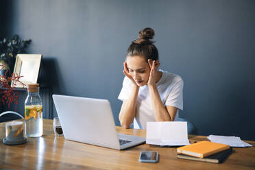 Female entrepreneur with head in hands sitting at desk against wall - BSZF01582