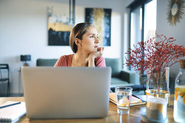 Thoughtful young woman with laptop on desk looking away while sitting at home - BSZF01596