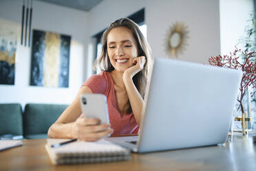 Smiling female freelancer using smart phone by laptop on desk in home office - BSZF01598