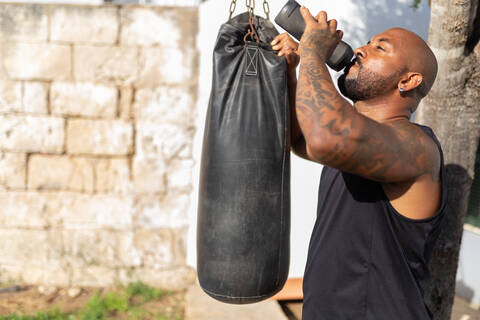 Bald mature man drinking water while standing by punching bag in yard stock photo