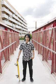 Junger Mann, der ein Skateboard hält, während er auf einer Fußgängerbrücke in der Stadt steht - XLGF00390