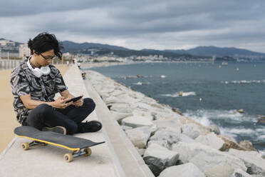 Junger Mann benutzt sein Smartphone, während er mit seinem Skateboard auf einer Stützmauer am Meer sitzt - XLGF00396