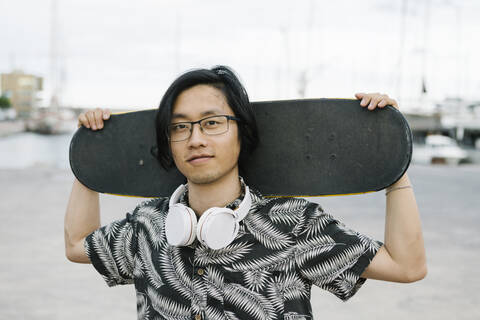 Nahaufnahme eines jungen Mannes mit Kopfhörern, der ein Skateboard hält und in der Stadt steht, lizenzfreies Stockfoto