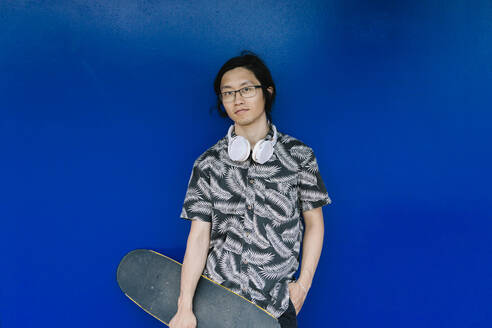 Junger Mann mit Skateboard an einer blauen Wand stehend - XLGF00401