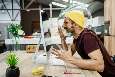 Frustrierter Geschäftsmann mit Fischglas im Kopf, der schreit, während er einen Computer in einem modernen Büro benutzt - RCPF00270