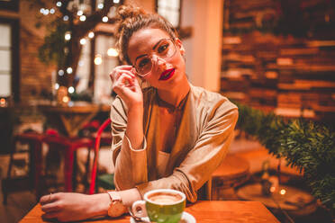 Attraktive junge Frau, die ihre Brille anfasst und in die Kamera schaut, während sie in einem Café mit einer Tasse Kaffee sitzt - ADSF05081
