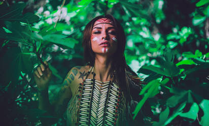 Attraktive junge Frau mit traditionellen indischen Malereien im Gesicht, die in die Kamera schaut und im Wald steht - ADSF05082
