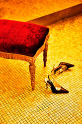Schwarze Lederschuhe mit hohen Absätzen auf dem Fliesenboden neben einem alten Stuhl mit gemusterten Beinen und rotem Bezug - ADSF05110