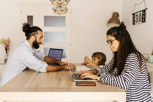 Frau arbeitet am Laptop, während Vater und Tochter zu Hause am Esstisch malen - JRFF04584