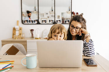 Smiling mother with daughter using laptop on dining table at home - JRFF04593