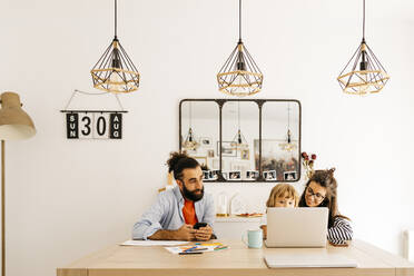 Man with smart phone looking at mother and daughter using laptop on dining table - JRFF04594