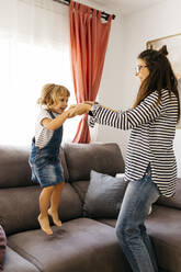 Glückliche Mutter, die die Hände ihrer verspielten Tochter hält, die zu Hause auf dem Sofa hüpft - JRFF04597