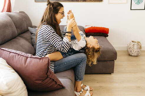 Mutter mit offenem Mund, die die Hände ihrer verspielten Tochter hält, während sie mit ihr auf dem Sofa zu Hause sitzt - JRFF04600