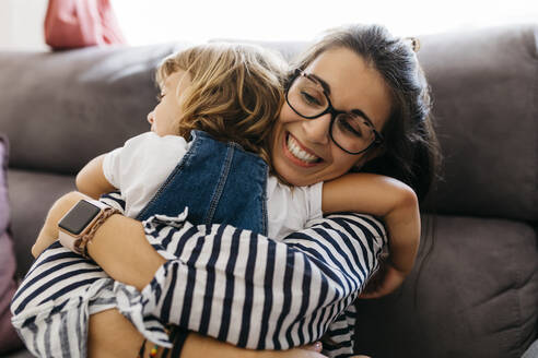 Nahaufnahme einer glücklichen Mutter, die ihre Tochter umarmt, während sie zu Hause auf dem Sofa sitzt - JRFF04603