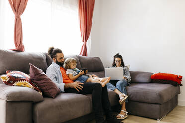 Frau arbeitet am Laptop, während Vater und Tochter auf dem Sofa zu Hause ein Smartphone benutzen - JRFF04604