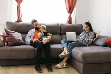 Frau arbeitet am Laptop, während Vater und Tochter auf dem Sofa im Wohnzimmer ein Mobiltelefon benutzen - JRFF04605