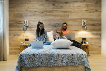 Parents with laptops looking at daughter playing with cushion on bed - JRFF04612