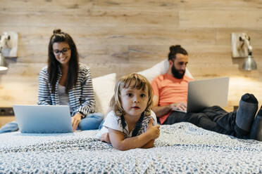 Nettes Mädchen auf dem Bett liegend, während die Mutter am Laptop arbeitet und der Vater im Schlafzimmer eingeschlafen ist - JRFF04615