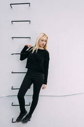 Junge hübsche blonde Frau hält sich an einer Leiter an der weißen Wand fest. - ADSF05319