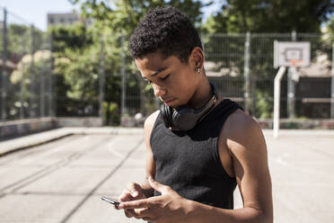 Afro-Junge hört Musik mit Smartphone und Kopfhörern auf dem Basketballplatz in seiner Nachbarschaft, Porträt - ADSF05396