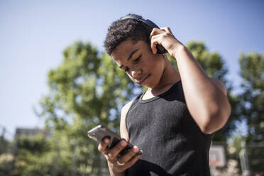 Afro-Junge hört Musik mit Smartphone und Kopfhörern auf dem Basketballplatz in seiner Nachbarschaft, Porträt - ADSF05398