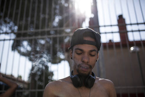 Afro-Junge hört Musik mit seinem Smartphone und Kopfhörern, während er auf dem Basketballplatz seines Viertels raucht und Rauch abgibt, Porträt - ADSF05399