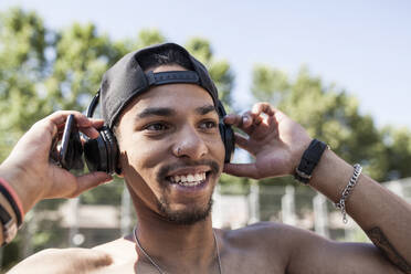 Afro Junge hört Musik mit seinem Smartphone und seinen Kopfhörern auf dem Basketballplatz in seiner Nachbarschaft, er lächelt, Portrait - ADSF05401