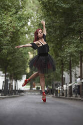 Red head ballerina with black tutu and red ballet tips dancing on the street with trees in the background. - ADSF05441