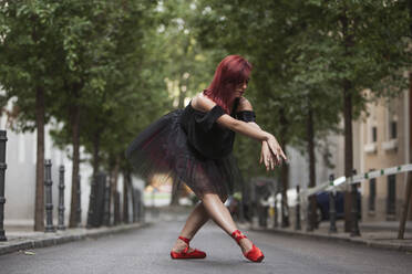 Red head ballerina with black tutu and red ballet tips dancing on the street with trees in the background. - ADSF05443