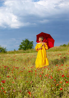 Ältere Frau mit rotem Regenschirm inmitten von Mohnblumen gegen den Himmel stehend - BFRF02266