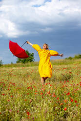 Unbekümmerte Frau mit ausgestreckten Armen, die einen roten Regenschirm hält, während sie in einem Mohnfeld steht - BFRF02267