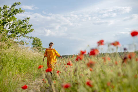 Happy mature woman walking amidst poppy field against sky stock photo