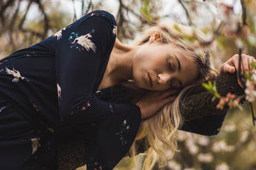 Junge blonde Frau lehnt sich an einen blühenden Baum und entspannt sich mit geschlossenen Augen in der Natur. - ADSF05739