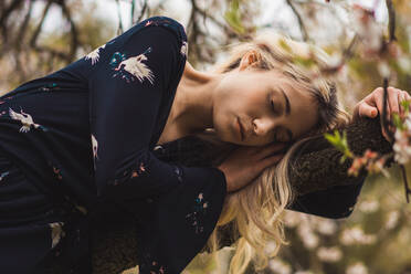 Young blonde woman leaning on blooming tree and relaxing with eyes closed in the nature. - ADSF05739