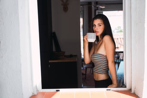 Seitenansicht einer jungen, schönen brünetten Frau in einem Crop-Top und einer schwarzen Strumpfhose, die in der Nähe des Fensters steht und Kaffee auf dem Hintergrund eines Raumes trinkt - ADSF05745