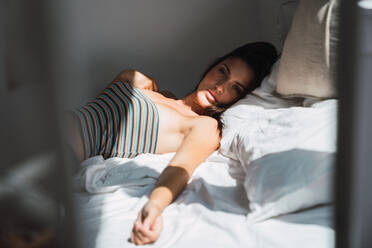 Side view of young beautiful woman with dark hair in striped crop top lying on white bed sheet in shadowed bedroom and looking at camera - ADSF05749