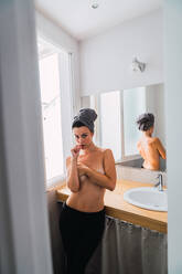 Junge attraktive barbusige Frau in schwarzer Strumpfhose und Handtuch auf dem Kopf, die im Badezimmer in der Nähe des Fensters steht und die Brust mit den Armen bedeckt - ADSF05752