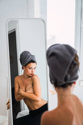 Rückenansicht einer jungen, schönen, barbusigen Frau mit einem grauen Handtuch auf dem Kopf, die in einem Raum vor einem Spiegel steht und ihre Brust mit den Armen bedeckt - ADSF05756