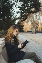 Seitenansicht einer hübschen Frau, die auf einer Parkbank sitzt und ein Buch liest. - ADSF05780