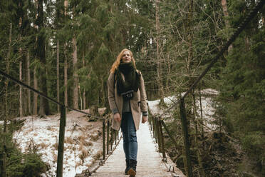 Frau in Wollmantel und Schal geht auf einem Steg im verschneiten Wald spazieren - FSIF05079